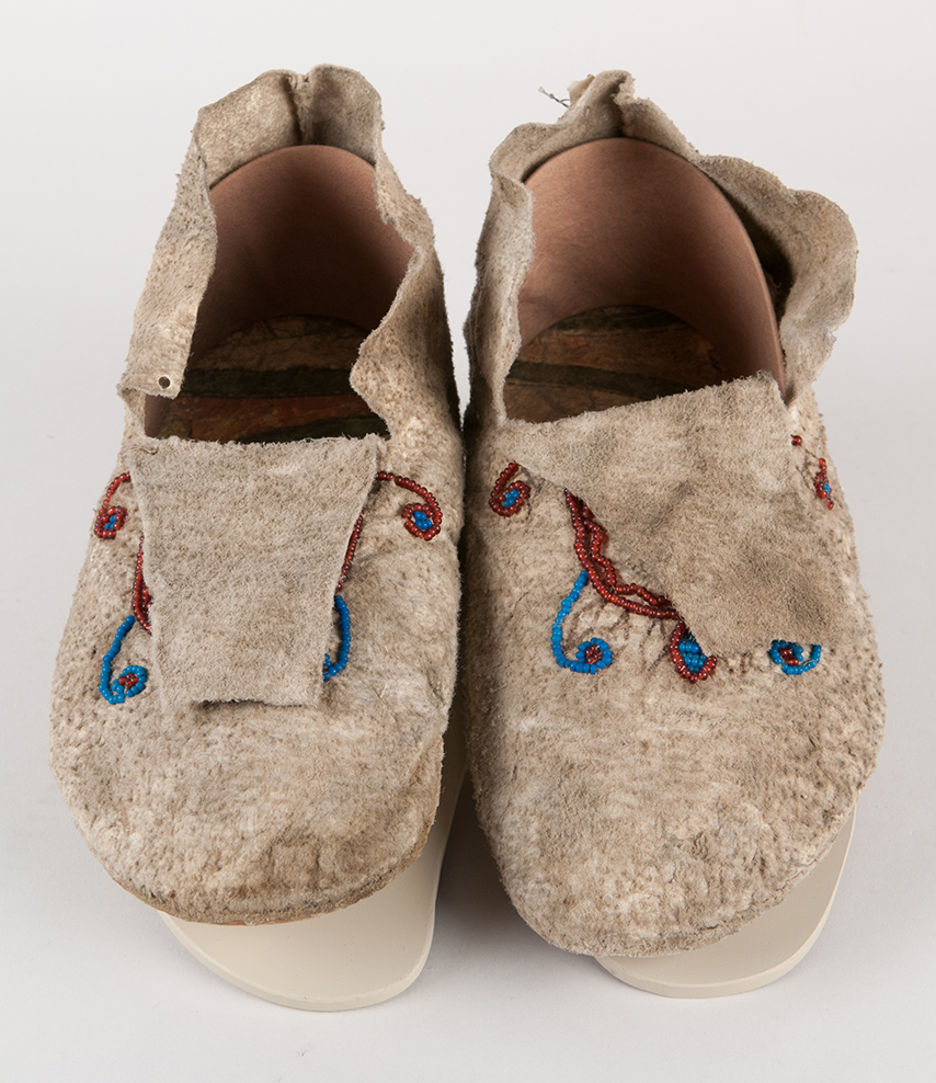 Pair of beaded moccasins made from three separate pieces of hide. The recycled rawhide features geometric painted designs of green, yellow, and red.